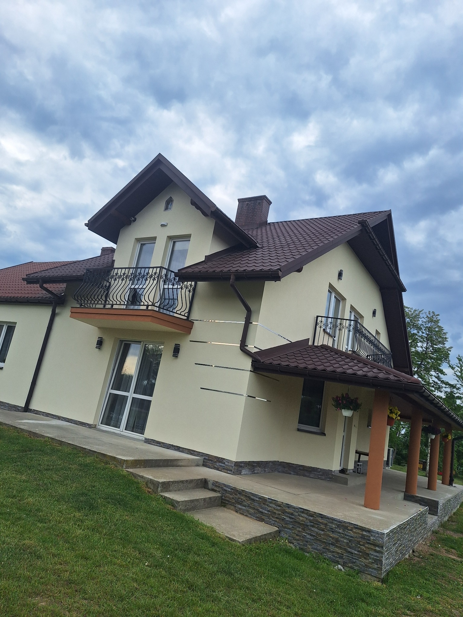 Elewacja domu jednorodzinnego w odcieniach beżu z brązowym dachem i balkonem z ozdobną balustradą, widok z poziomu trawnika przy pochmurnym niebie.