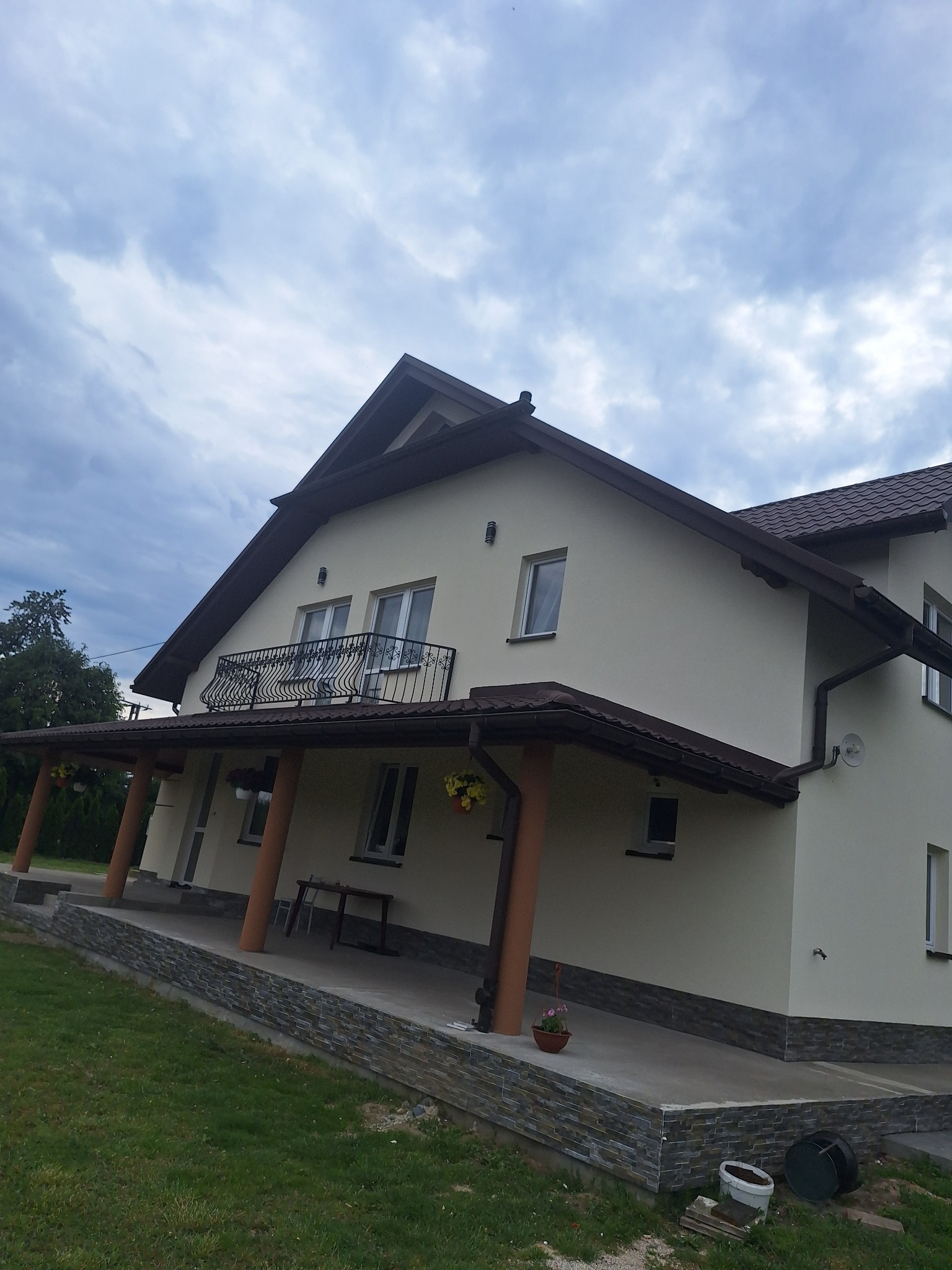 Dwukondygnacyjny dom z balkonem i zadaszonym tarasem, kremowa elewacja, brązowe elementy wykończeniowe, widok z dołu na tle pochmurnego nieba.