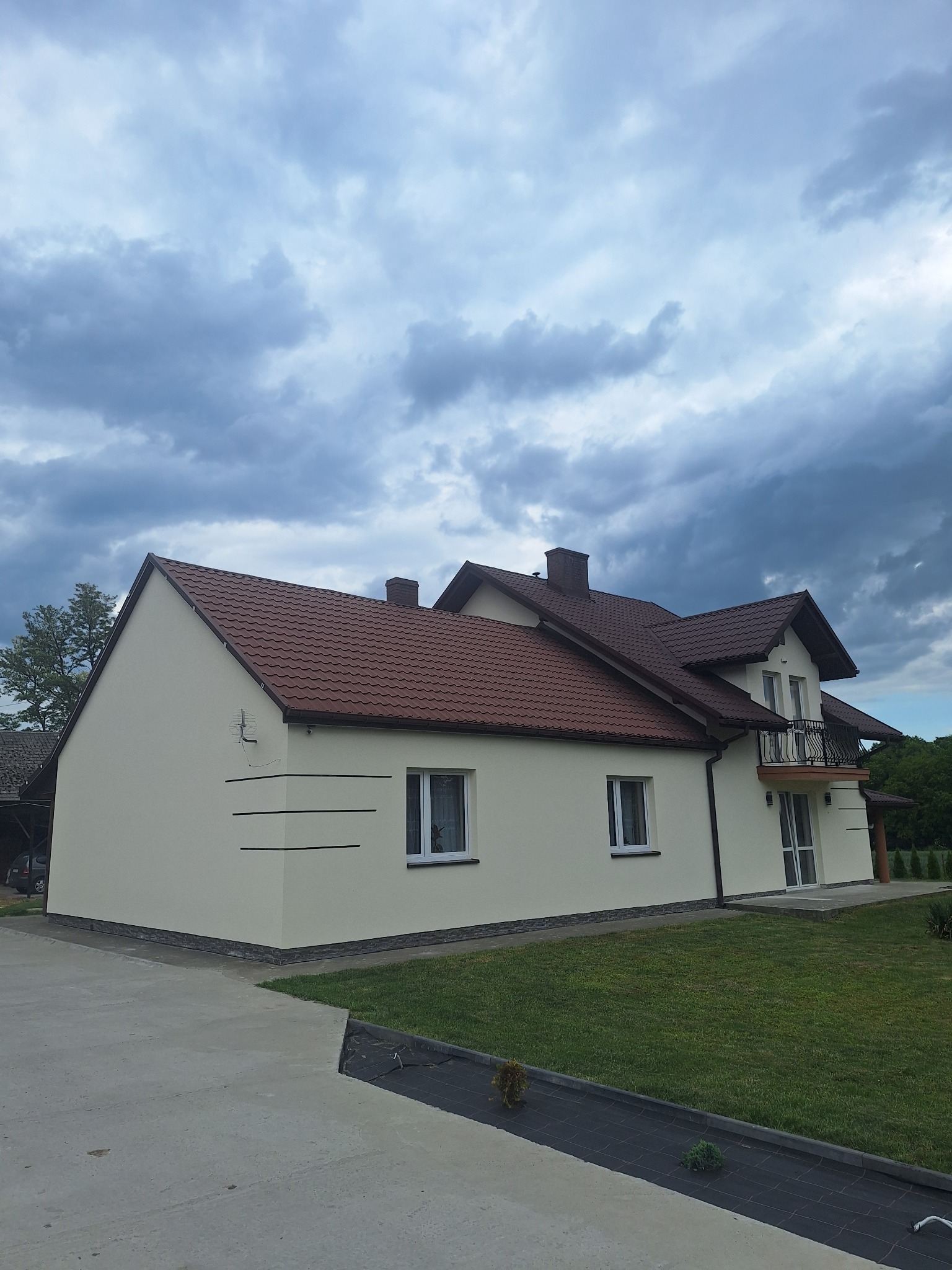 Beżowy dom z ciemną, czerwoną dachówką, balkonem i ozdobnymi poziomymi paskami na elewacji, widziany z poziomu trawnika w pochmurny dzień.