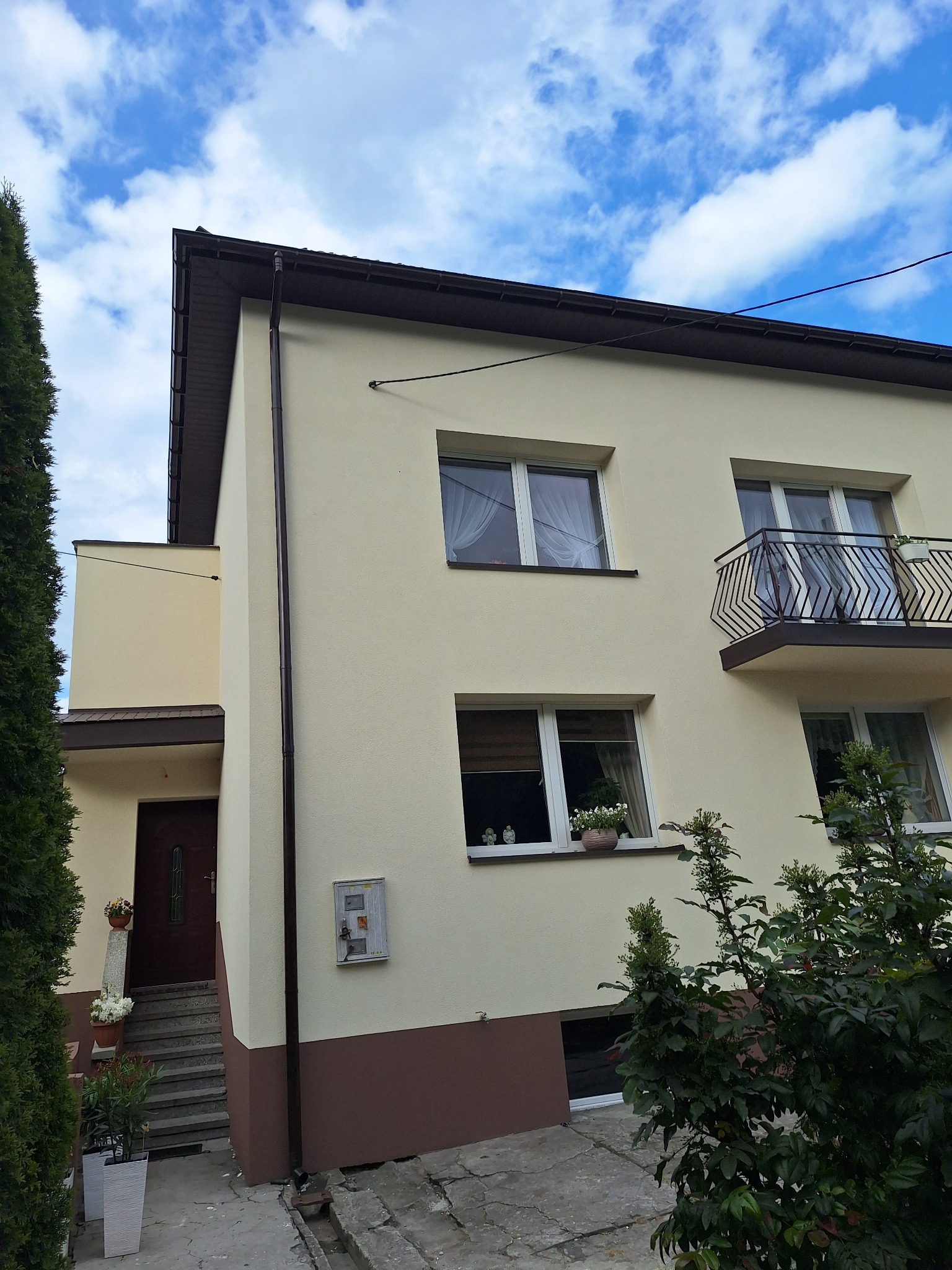 Dwukondygnacyjny dom z beżową elewacją i brązowym cokołem, balkon z ozdobną balustradą, okna z kwiatami, błękitne niebo w tle. Zwraca uwagę staranne wykończenie.