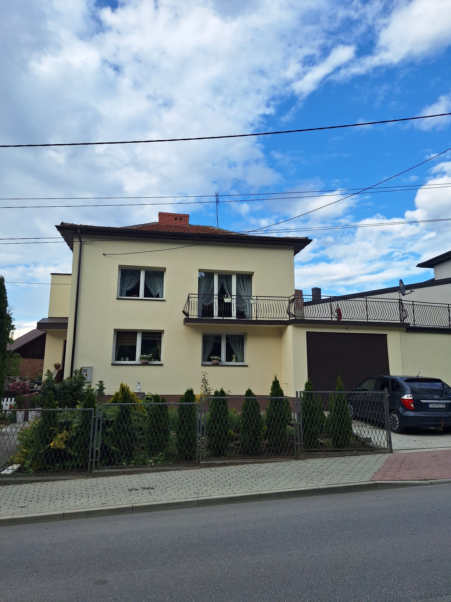Dwupiętrowy dom z beżową elewacją, balkonem z czarną balustradą i garażem. Na podjeździe stoi ciemny samochód. W tle błękitne niebo z chmurami.