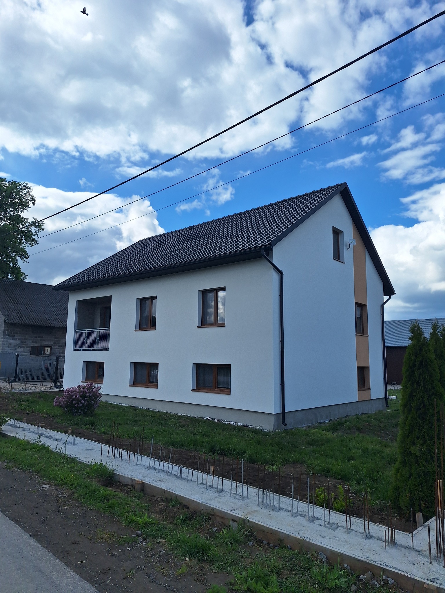 Dwukondygnacyjny dom z białą elewacją i ciemną dachówką w słoneczny dzień. Widoczne elementy dekoracyjne elewacji i przygotowane fundamenty pod ogrodzenie.