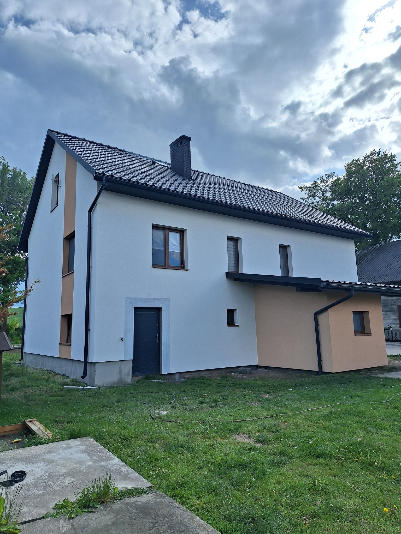 Dwukondygnacyjny dom z białą elewacją i brązowymi akcentami pod pochmurnym niebem. Widoczne okna, drzwi wejściowe i fragment dachu z kominem.