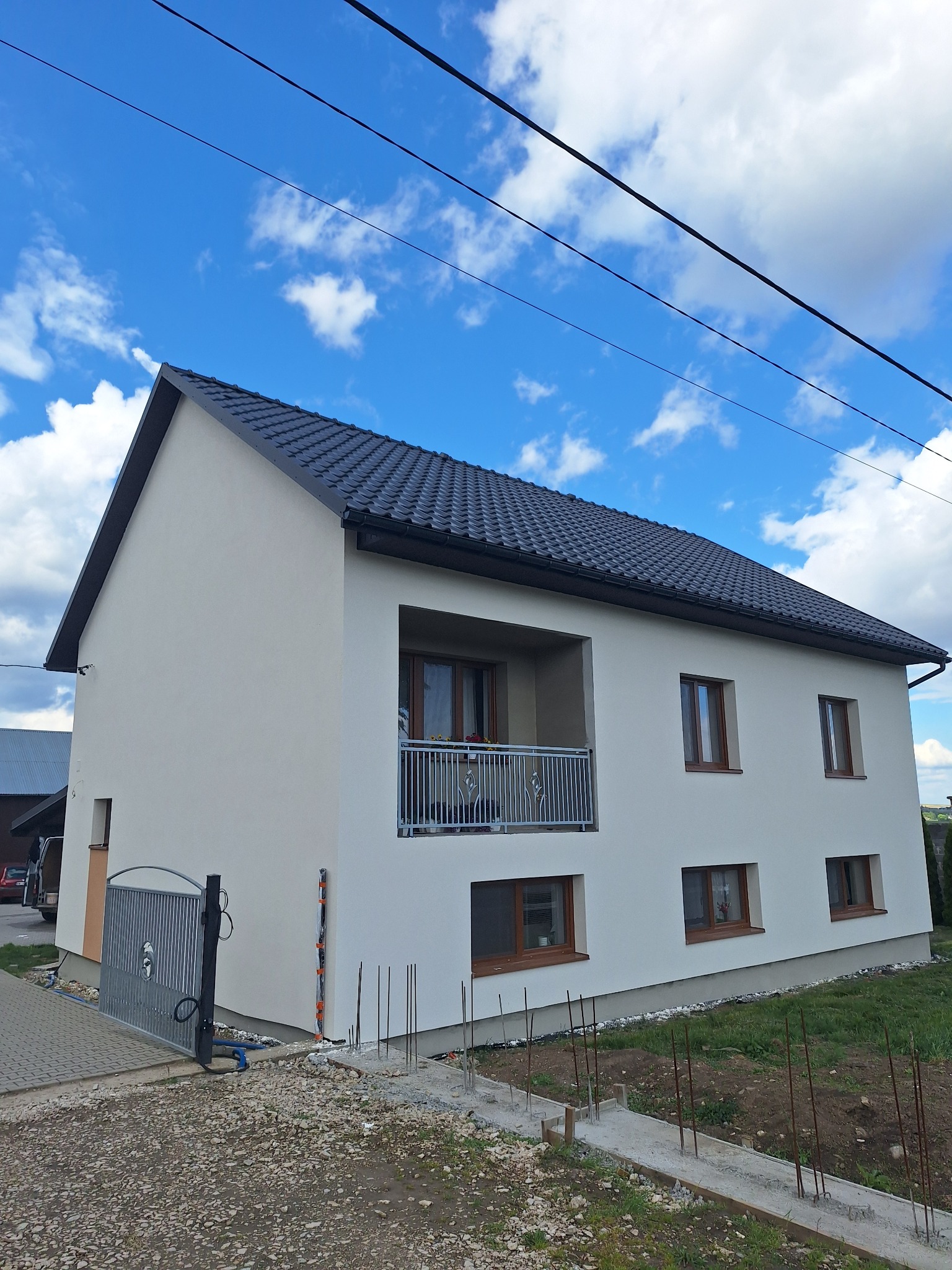 Dwukondygnacyjny dom z balkonem i szarą dachówką pod błękitnym niebem. Biała elewacja, brązowe okna. Widoczne elementy przygotowania pod dalsze prace.