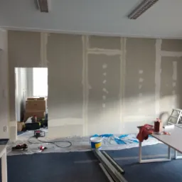 Wnętrze biurowe w trakcie prac wykończeniowych: ściany z płyt karton-gips, widoczne łączenia przygotowane do szpachlowania, zabezpieczona podłoga, drabina i narzędzia pracy.