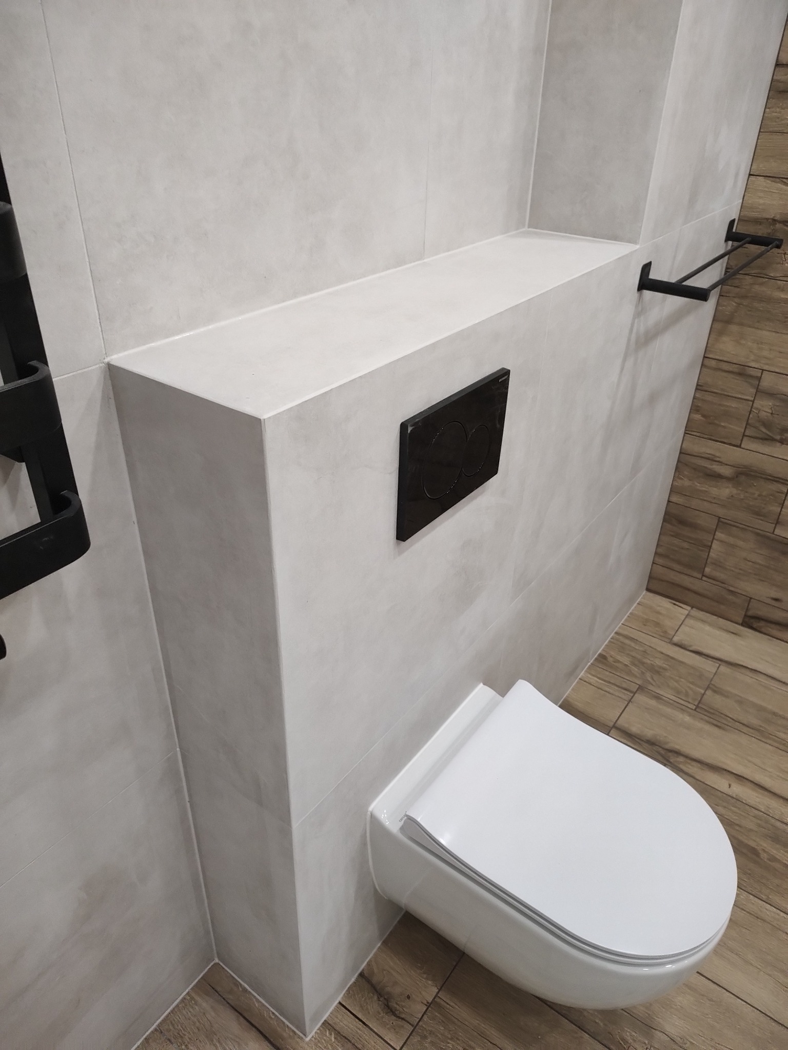 Nowoczesna toaleta podwieszana z czarnym przyciskiem spłukującym, obudowana płytkami w odcieniach szarości, z drewnopodobną podłogą i czarnym grzejnikiem drabinkowym.