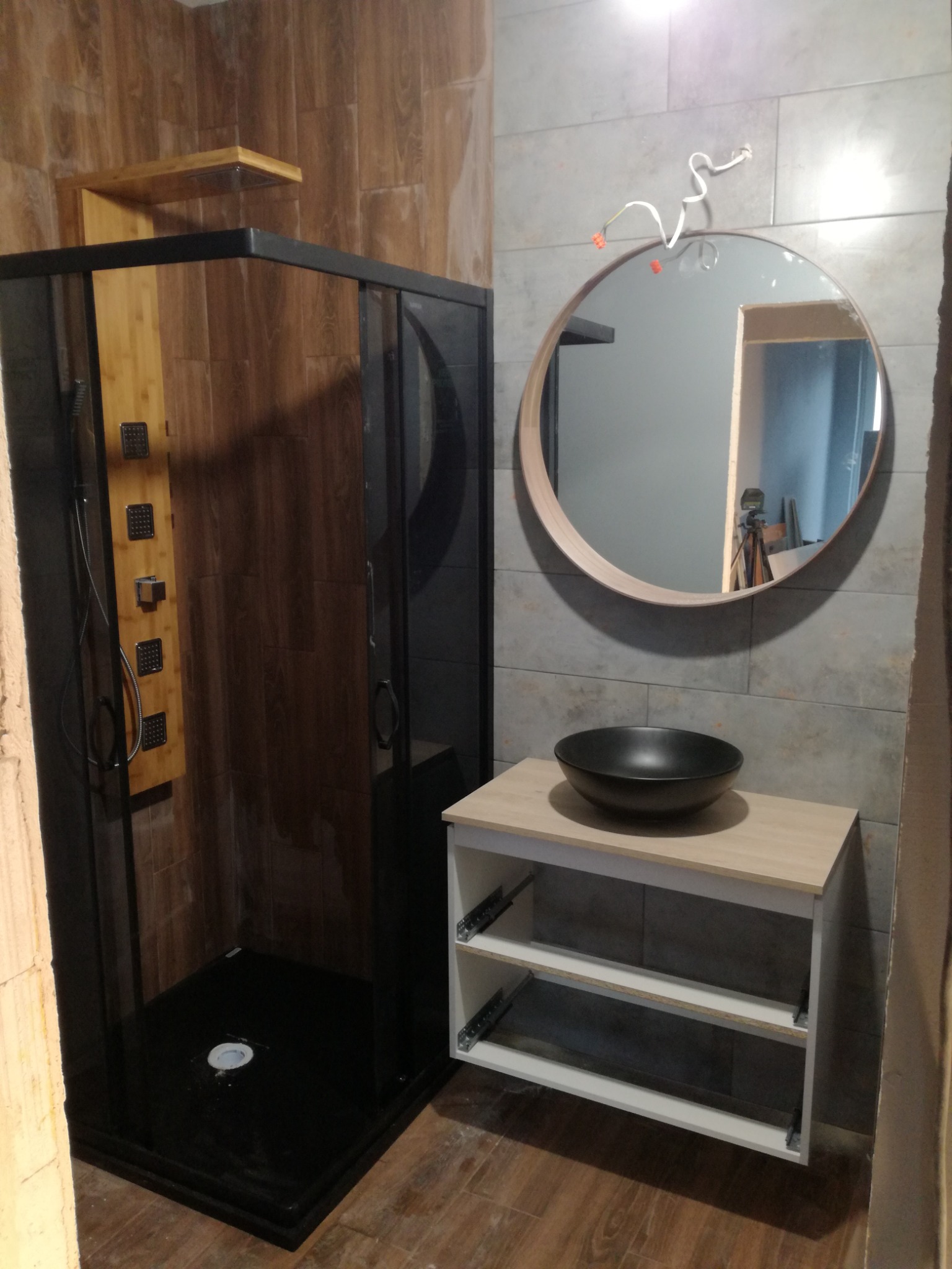 Nowoczesna łazienka z czarną kabiną prysznicową typu walk-in, drewnopodobnymi płytkami na ścianach oraz okrągłym lustrem z drewnianą ramą nad umywalką nablatową.