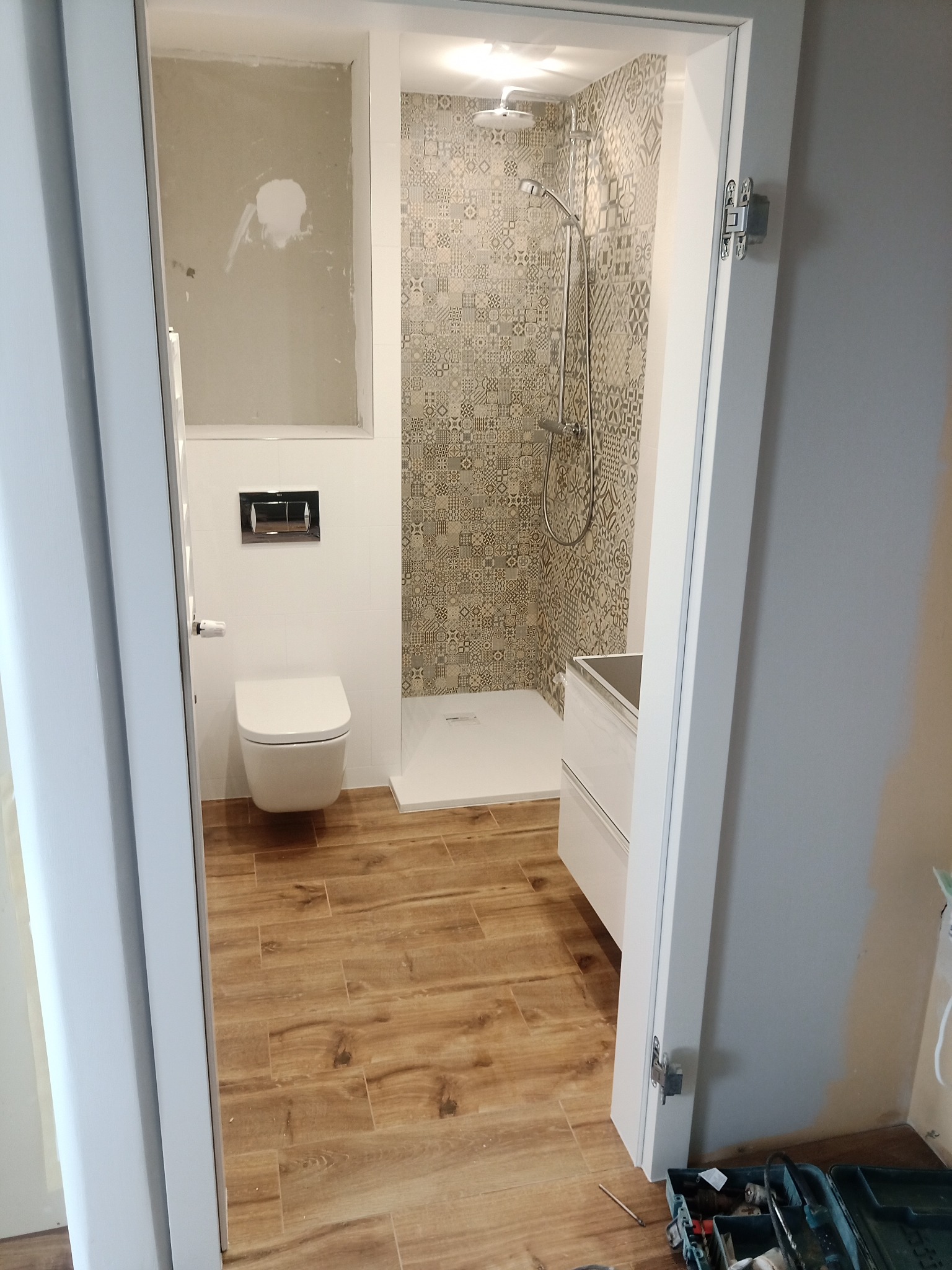 Nowoczesna łazienka w trakcie remontu, widoczny fragment ściany przygotowanej pod płytki, zamontowana toaleta podwieszana, kabina prysznicowa z wzorzystymi płytkami i drewnopodobna podłoga.