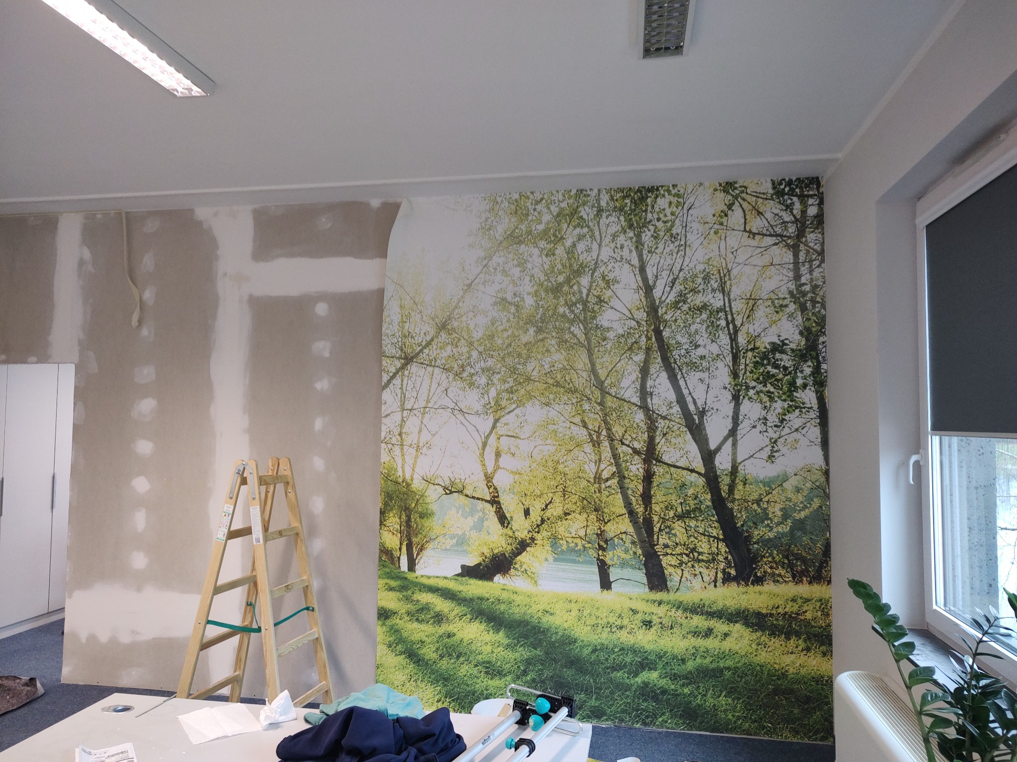 Ściana w trakcie remontu z częściowo nałożoną fototapetą z motywem leśnym, widoczna drabina i narzędzia na stole.