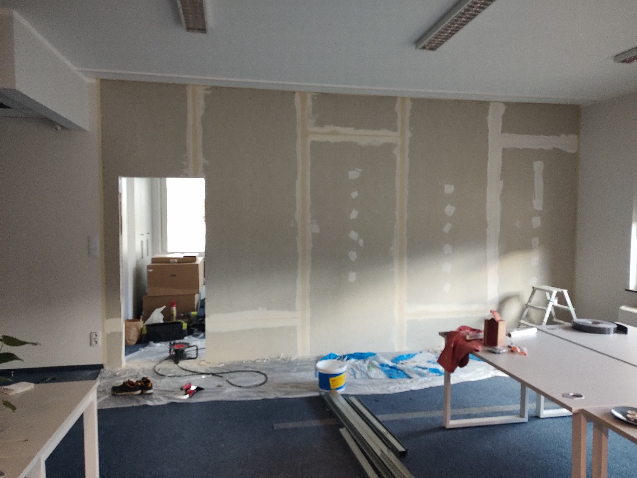 Wnętrze biurowe w trakcie prac wykończeniowych: ściany z płyt karton-gips, widoczne łączenia przygotowane do szpachlowania, zabezpieczona podłoga, drabina i narzędzia pracy.