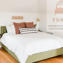 Tapicerowane łóżko z zieloną ramą i białym obiciem, ozdobione brązowymi i czarno-białymi poduszkami, ustawione w jasnym pokoju z drewnianą podłogą i złotymi lampami ściennymi, obok mały stolik...