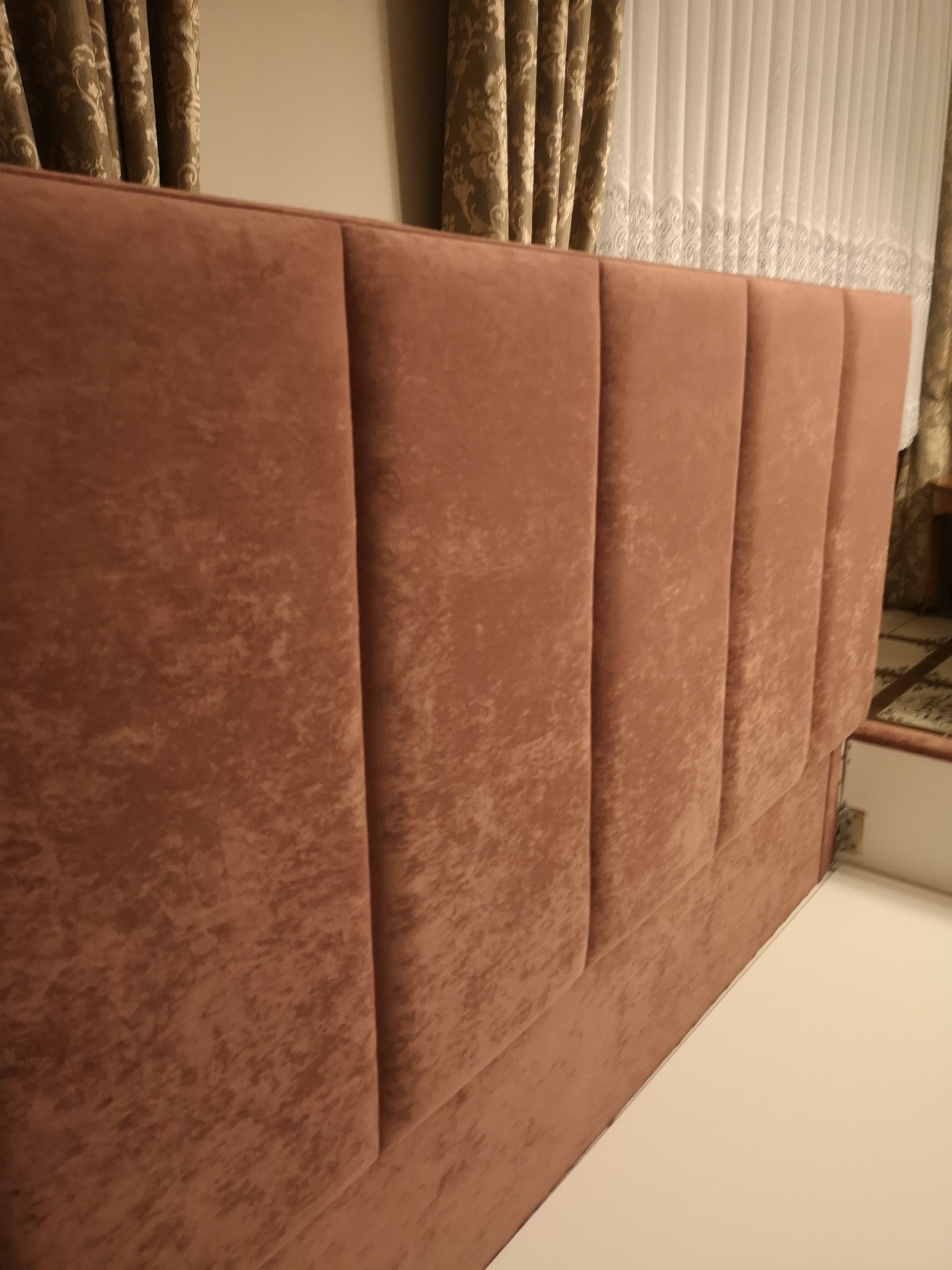 Wezgłowie łóżka tapicerowane brązowym welurem, podzielone na pionowe panele z widoczną strukturą materiału, na tle zasłon w odcieniach brązu i bieli.