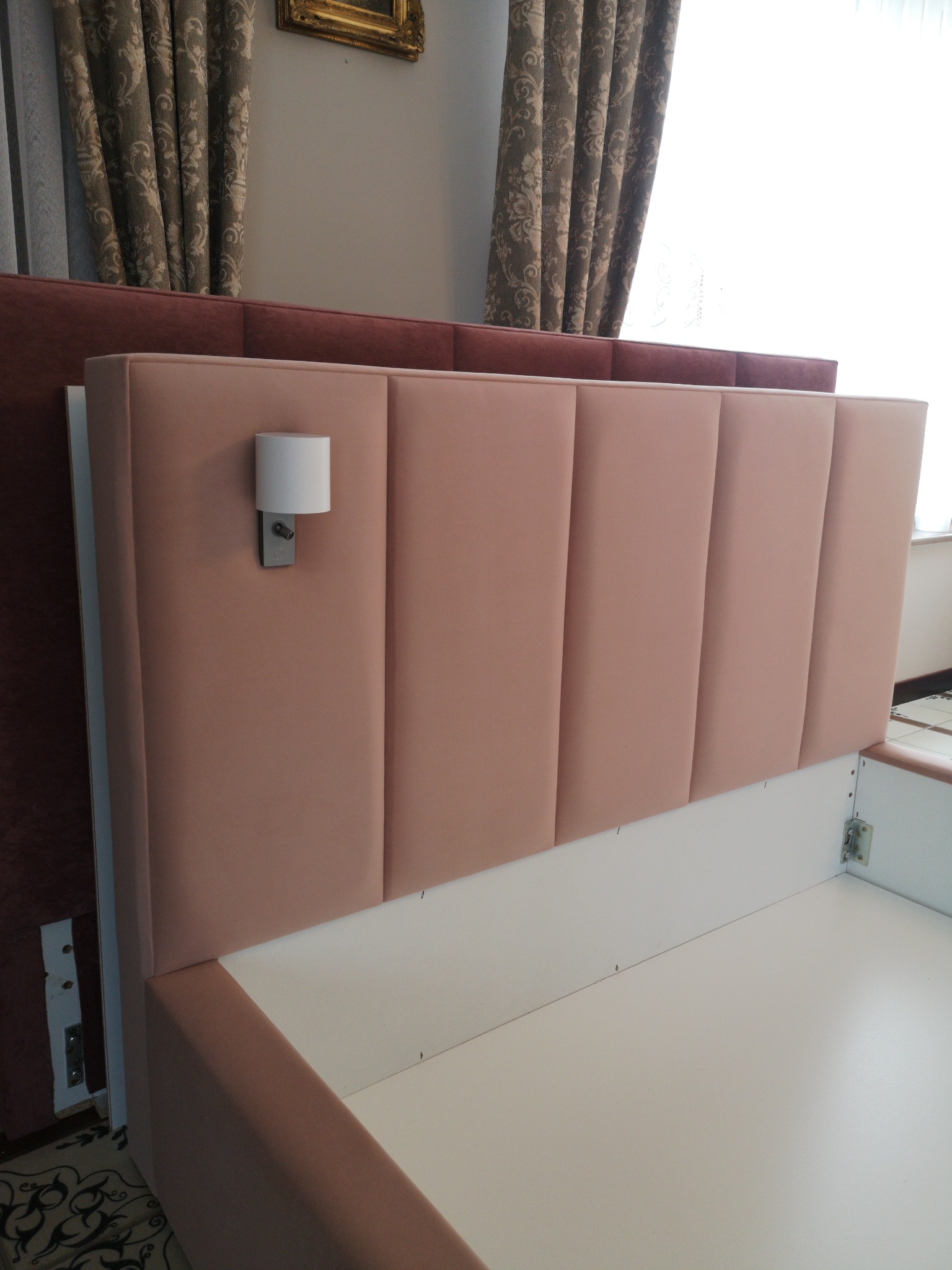 Różowa, tapicerowana rama łóżka z pionowymi panelami i zamontowaną lampką nocną, widoczne elementy montażowe i fragment podłogi z dywanem.