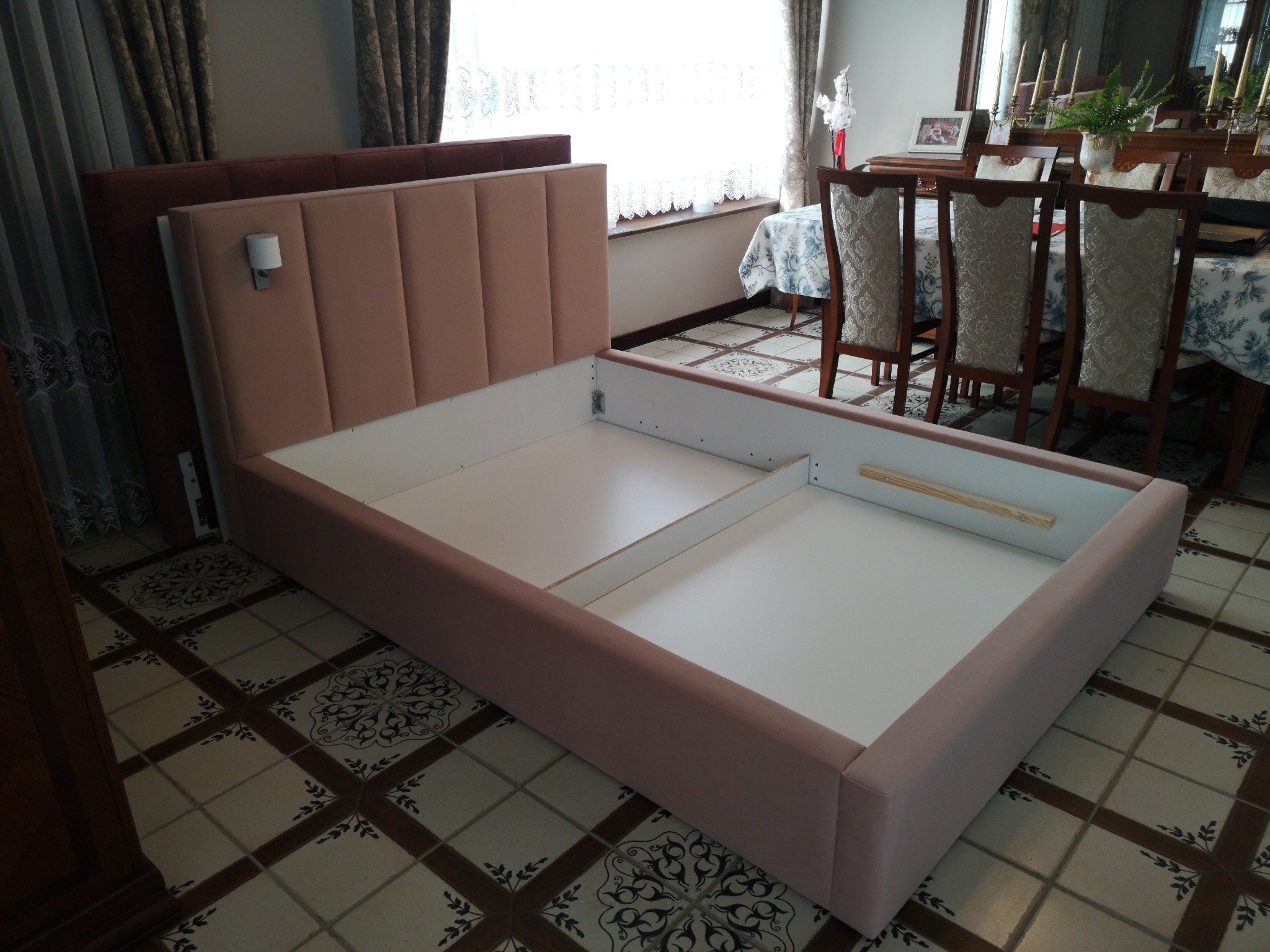 Różowa tapicerowana rama łóżka z pionowymi przeszyciami na wezgłowiu, widoczna w trakcie montażu, z elementami mocującymi, na tle podłogi z wzorzystych płytek.