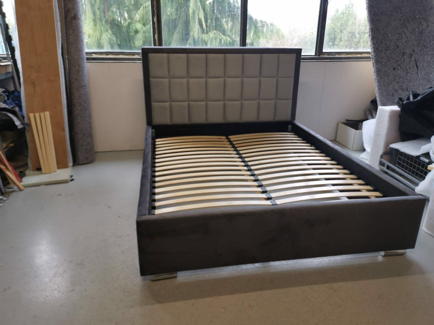 Tapicerowane łóżko z szarym zagłówkiem w kratkę i drewnianym stelażem, na srebrnych nóżkach, w pomieszczeniu z oknem i materiałami tapicerskimi.