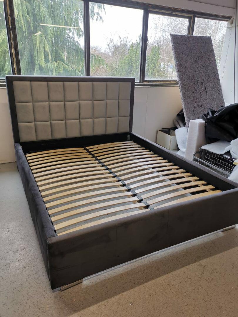 Tapicerowane łóżko z szarym, pikowanym wezgłowiem i drewnianym stelażem, widoczne na tle okna i elementów magazynowych.