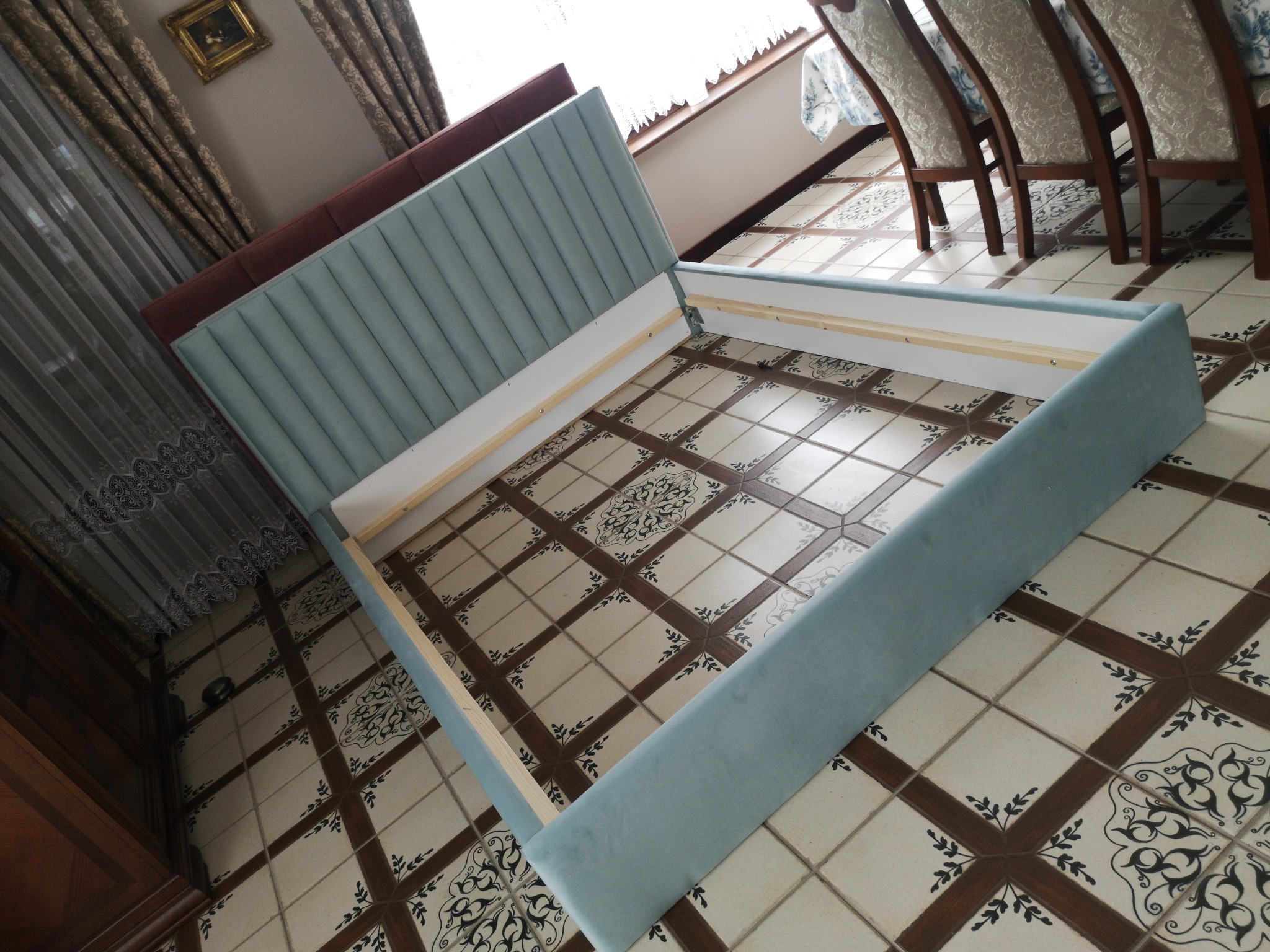 Narożne ujęcie ramy łóżka z jasnoniebieskim tapicerowanym wezgłowiem i bokami, spoczywającej na ozdobnej, wzorzystej podłodze z płytek.
