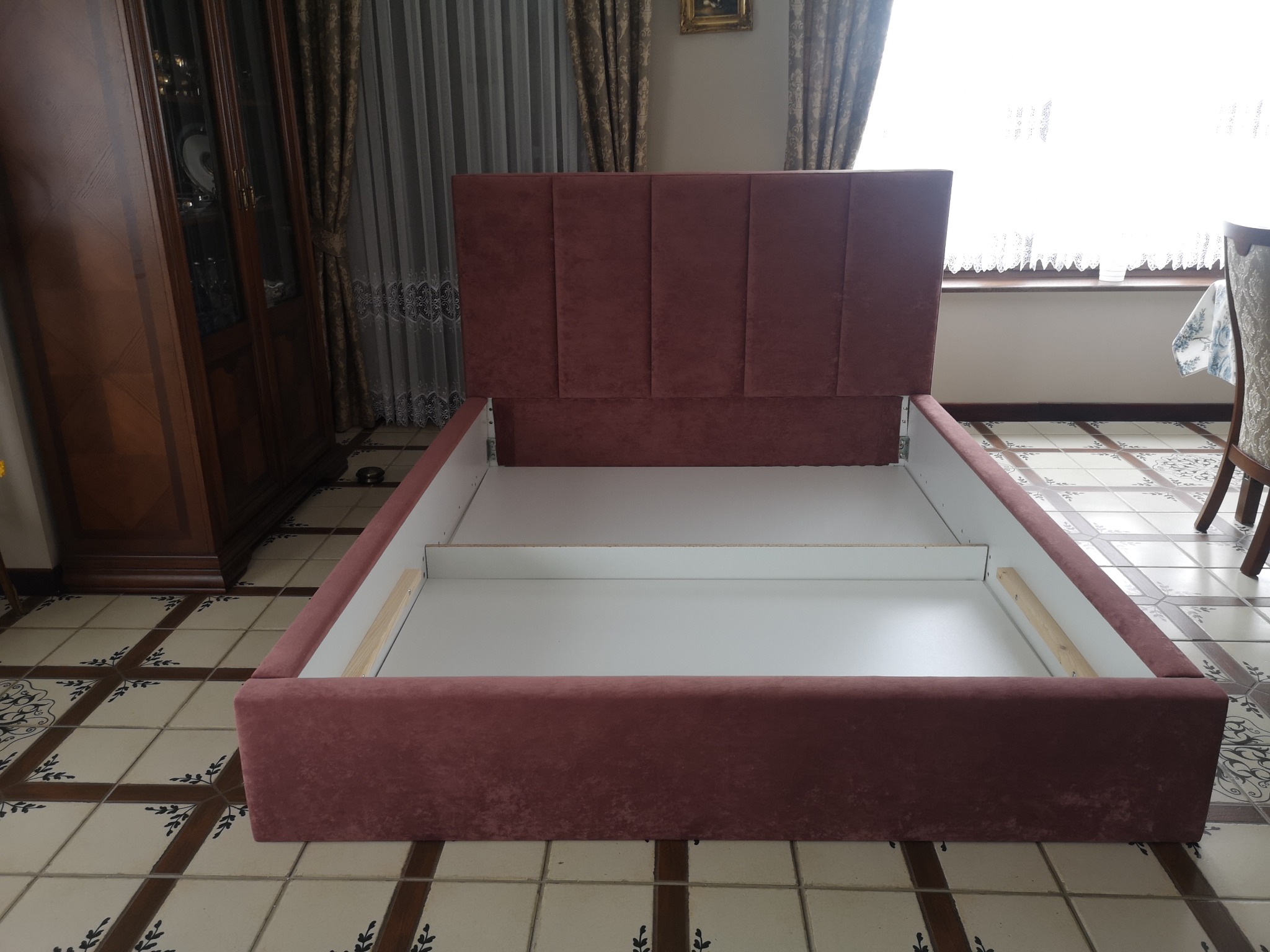 Rozłożone łóżko z bordową, tapicerowaną ramą i zagłówkiem, widoczne białe wnętrze ramy i drewniane elementy montażowe, na tle wzorzystej podłogi i mebli.