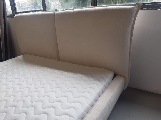 Tapicerowane beżową tkaniną łóżko z wysokim zagłówkiem i wzorzystym materacem, ustawione pod oknem z widocznymi zasłonami.