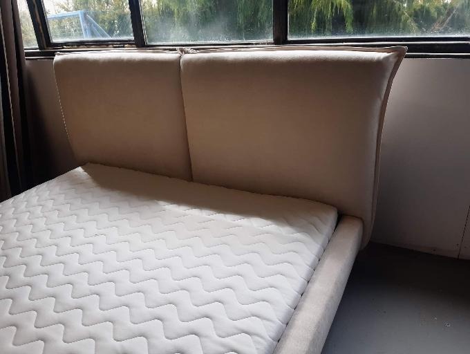 Beżowe tapicerowane łóżko z zagłówkiem, widok z boku, materac w białym pokrowcu ze wzorem, okno w tle.