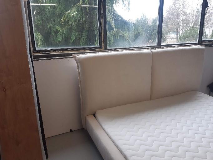 Beżowe, tapicerowane wezgłowie łóżka z materacem we Wrocławiu, widoczne na tle dużego okna z widokiem na drzewa.