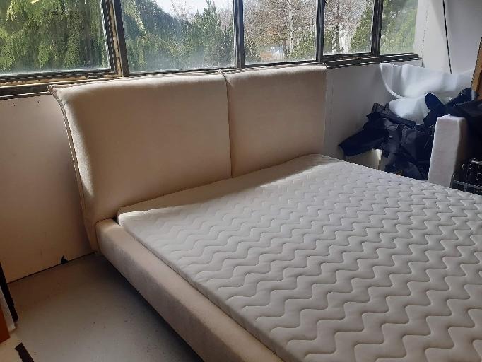 Beżowe tapicerowane łóżko z materacem w pokoju z widokiem na zieleń za oknem, widoczne niedoskonałości wykończenia i bałagan w tle.