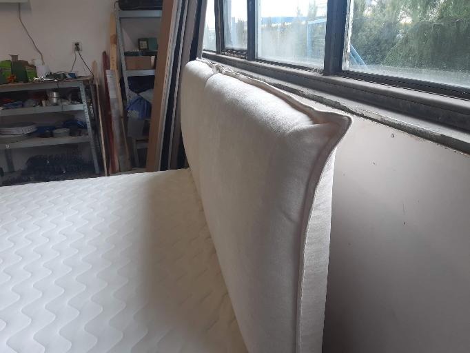 Białe, tapicerowane wezgłowie łóżka z widocznym szwem wykończeniowym, ustawione w warsztacie z oknem w tle.