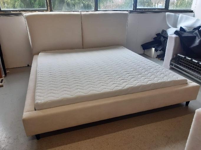 Beżowe łóżko tapicerowane z zagłówkiem i materacem w surowym pomieszczeniu z dużym oknem, widoczne ślady pracy tapicerskiej.