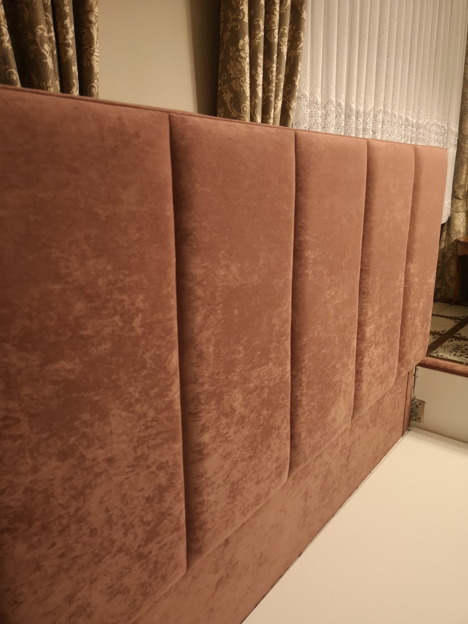 Wezgłowie łóżka obite brązowym welurem z pionowymi przeszyciami, widoczne z boku, na tle zasłon w podobnej tonacji.