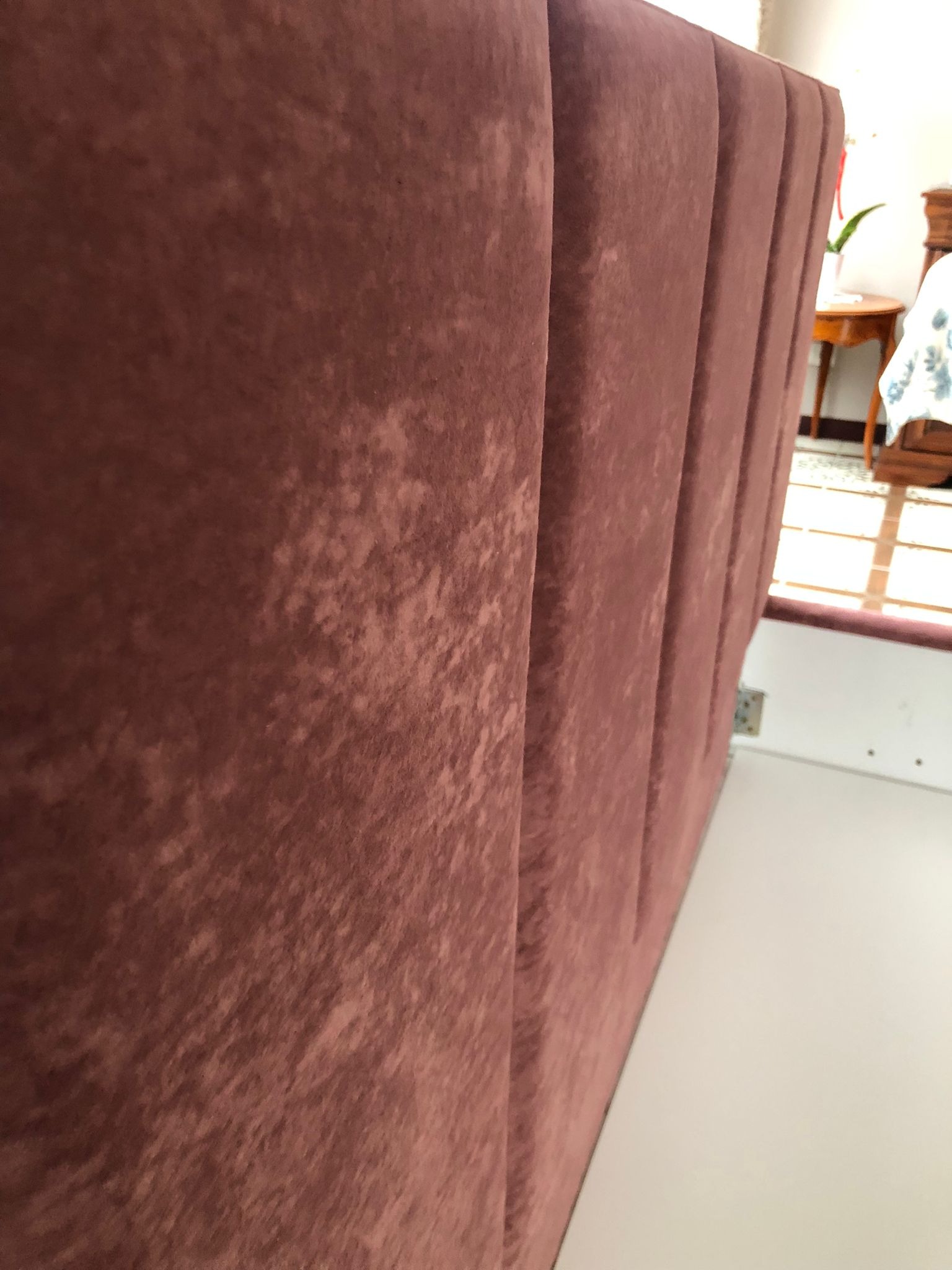 Szczegółowe ujęcie wezgłowia łóżka z bordowej tkaniny z pionowymi przeszyciami, widoczne delikatne cieniowanie materiału i fragment pokoju z drewnianym stolikiem w tle.