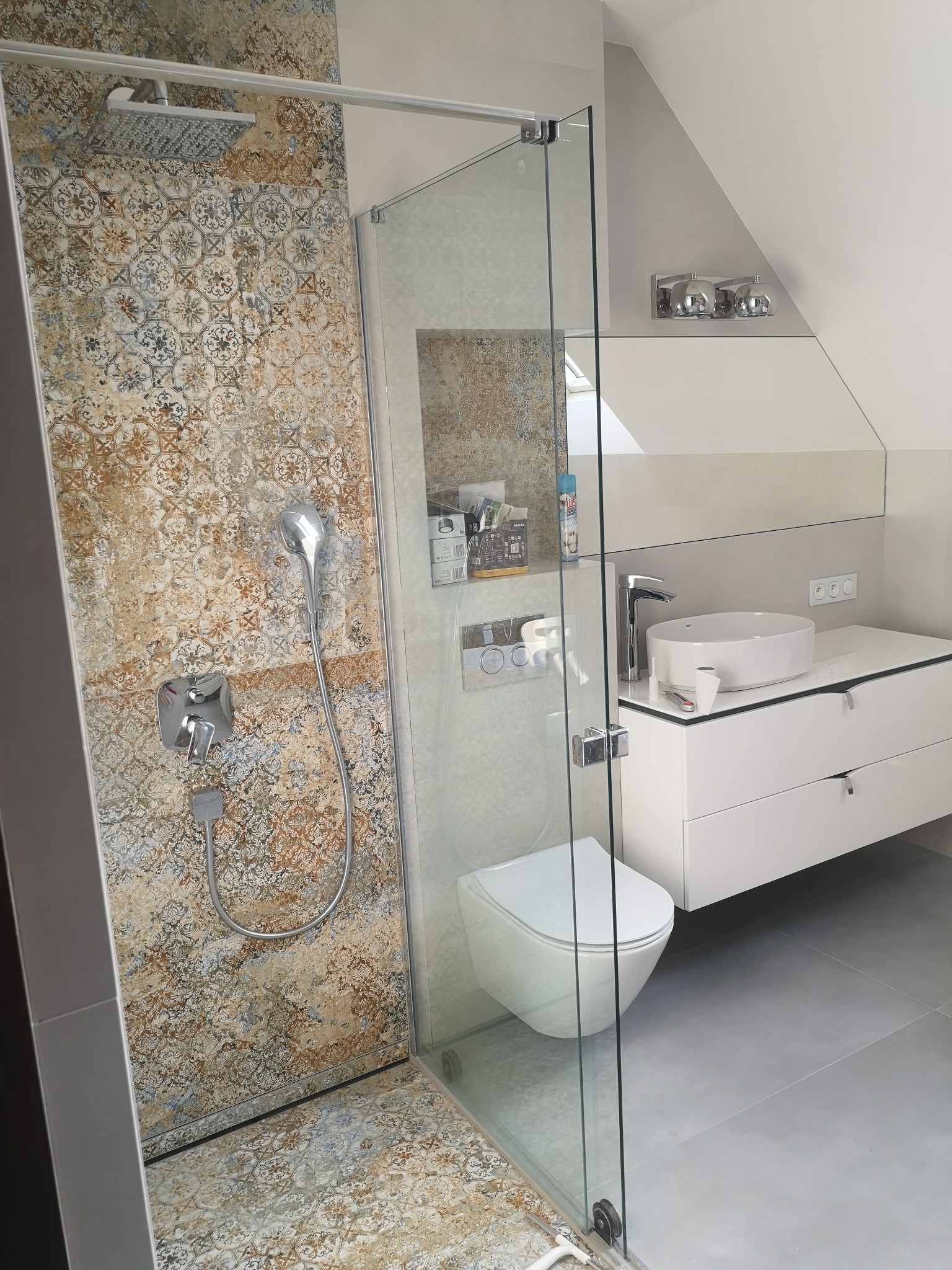 Nowoczesna łazienka na poddaszu z kabiną prysznicową typu walk-in, zdobionymi płytkami w strefie prysznica i minimalistyczną białą szafką z umywalką nablatową.