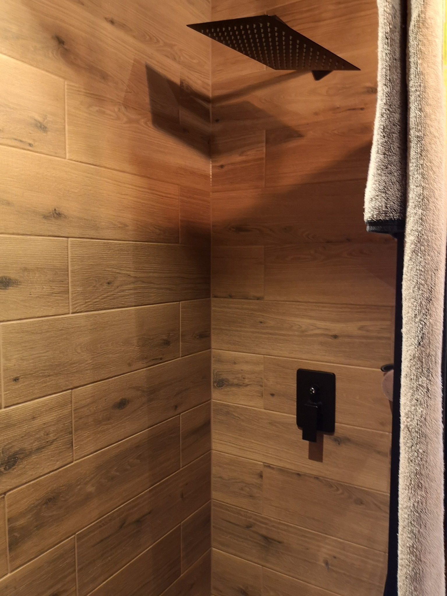 Kabina prysznicowa wyłożona płytkami imitującymi drewno, z czarną deszczownicą i baterią podtynkową oraz wiszącym ręcznikiem w kolorze szarym.