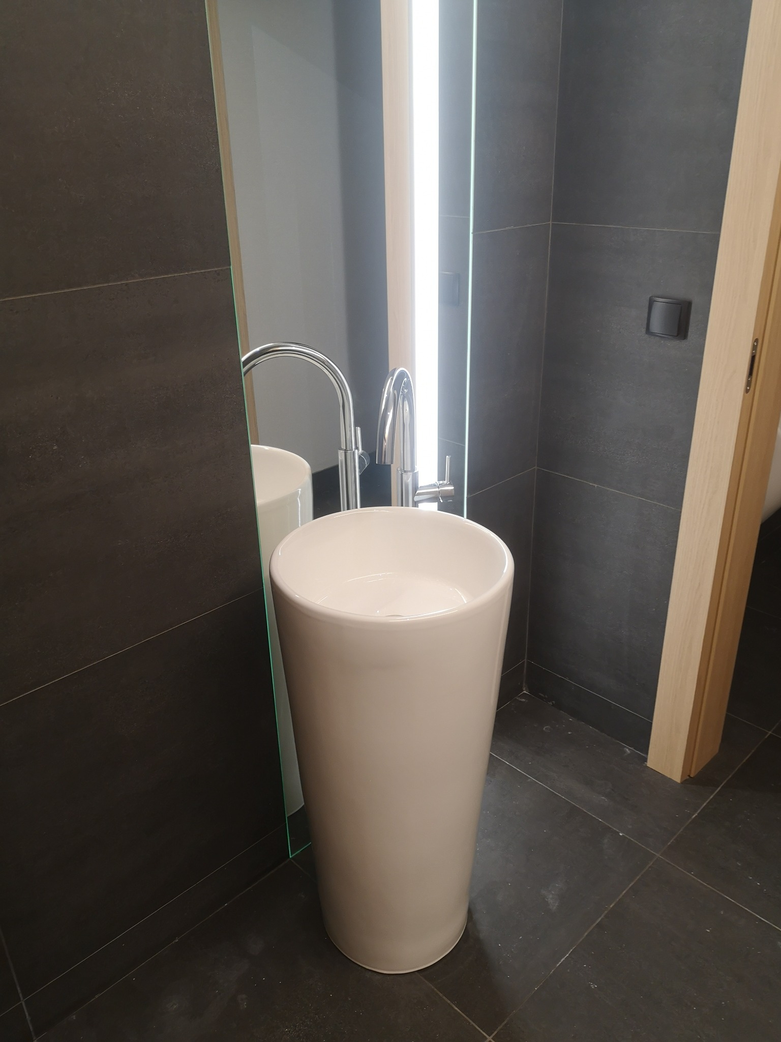 Biała, wolnostojąca umywalka z chromowaną baterią na tle ciemnych płytek w łazience z podświetlanym lustrem.