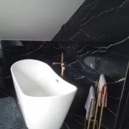 Elegancka, biała wanna wolnostojąca z złotą armaturą na tle czarnych marmurowych płytek w łazience w Kozy. Widoczny również złoty wieszak na ręczniki i szary dywanik.