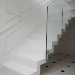 Nowoczesne białe schody z szklaną balustradą, mocowaną za pomocą metalowych uchwytów, w jasnym wnętrzu z dekoracyjną sztukaterią na ścianie.