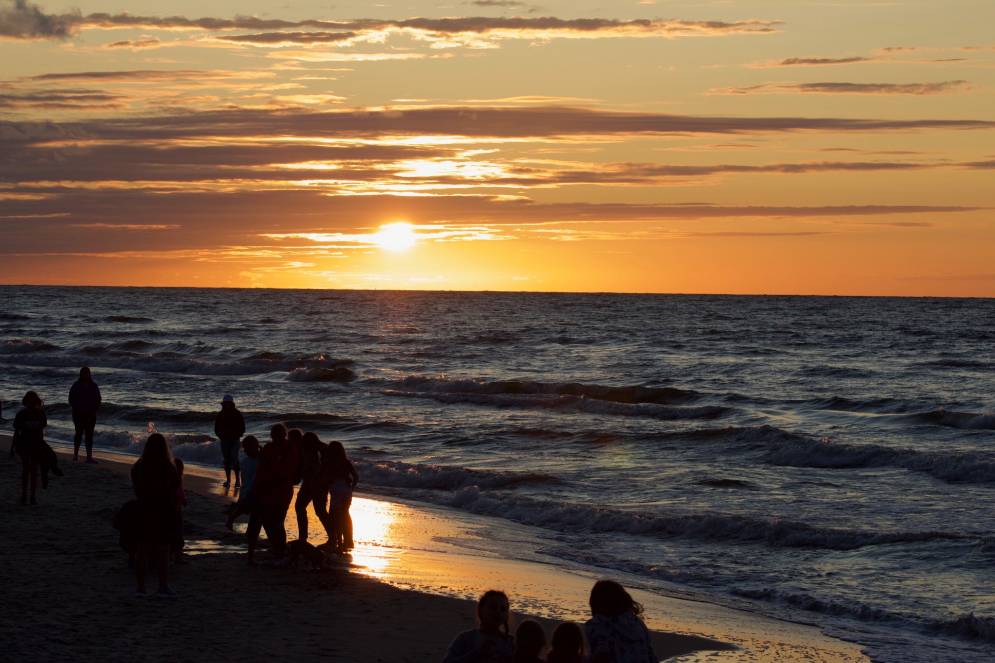Sylwetki grupy osób na plaży podczas zachodu słońca nad morzem, z refleksami światła na piasku i falach.
