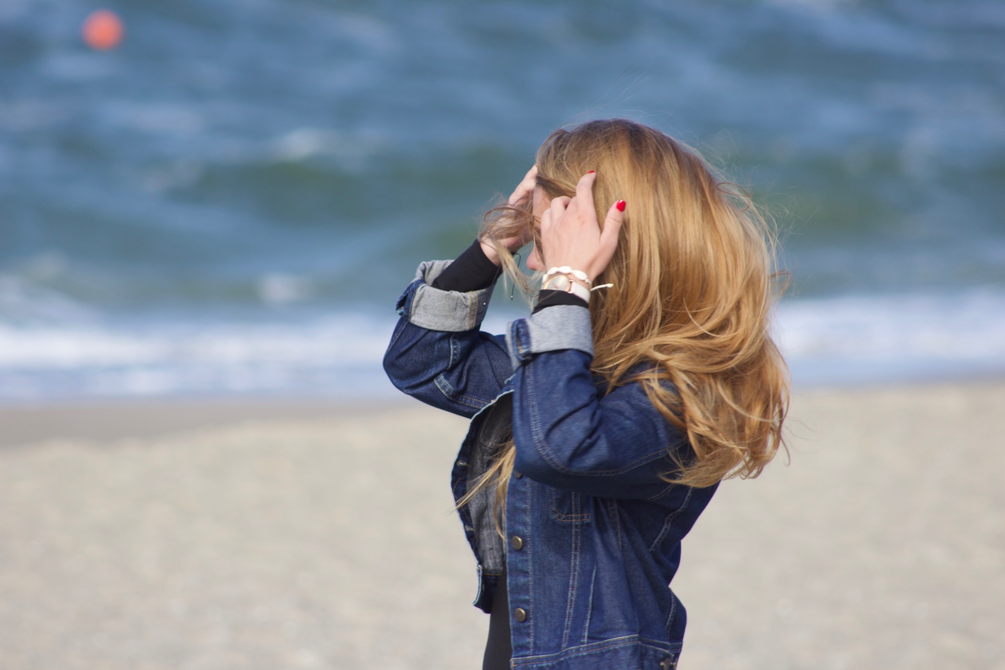 Kobieta w dżinsowej kurtce na plaży, poprawiająca włosy, z widokiem na morze i odległą czerwoną boję.