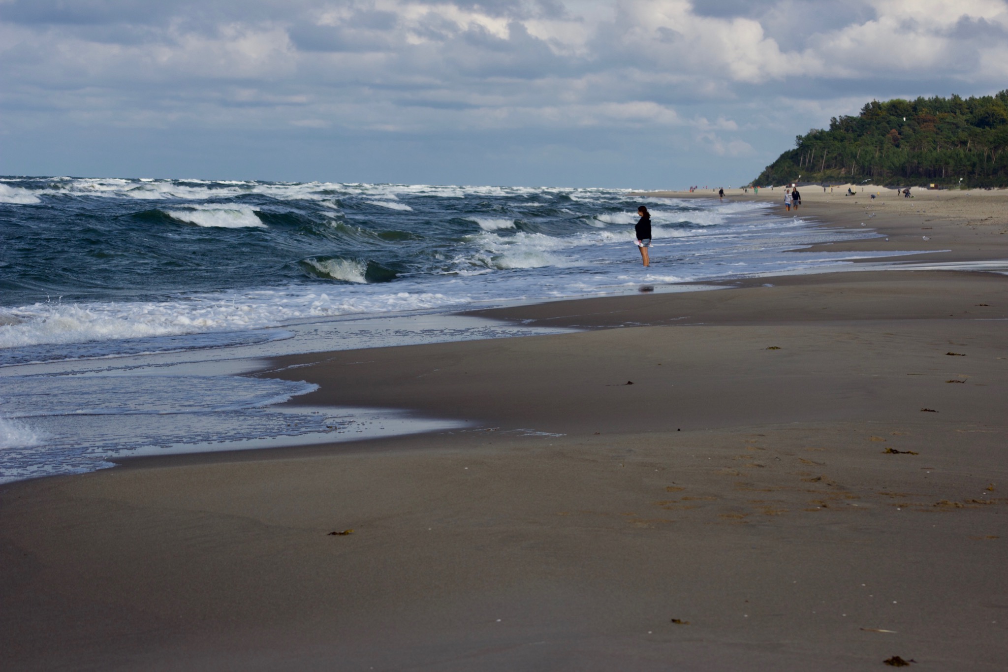 Kobieta stoi w płytkiej wodzie na piaszczystej plaży, obserwując wzburzone morze pod pochmurnym niebem. W tle widać drzewa i spacerujących ludzi.
