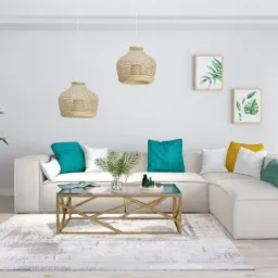 Jasny salon z narożną sofą, szklanym stolikiem kawowym na złotym stelażu, wiszącymi lampami z abażurami z naturalnego materiału, rośliną doniczkową w ozdobnej osłonce i grafikami z motywem...