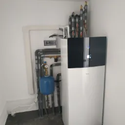 Nowoczesna pompa ciepła Daikin zamontowana w pomieszczeniu, widoczne rury instalacyjne, manometr, zabezpieczenia termiczne oraz skrzynka elektryczna na ścianie.