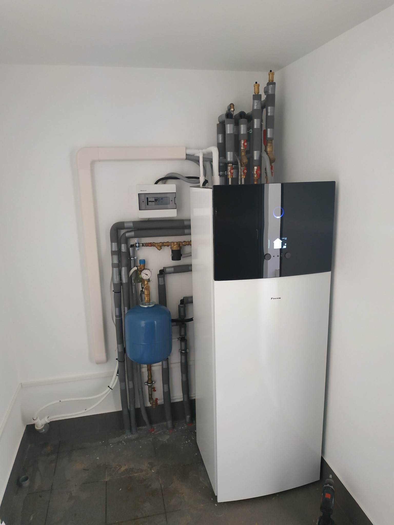 Nowoczesna pompa ciepła Daikin zamontowana w pomieszczeniu, widoczne rury instalacyjne, manometr, zabezpieczenia termiczne oraz skrzynka elektryczna na ścianie.