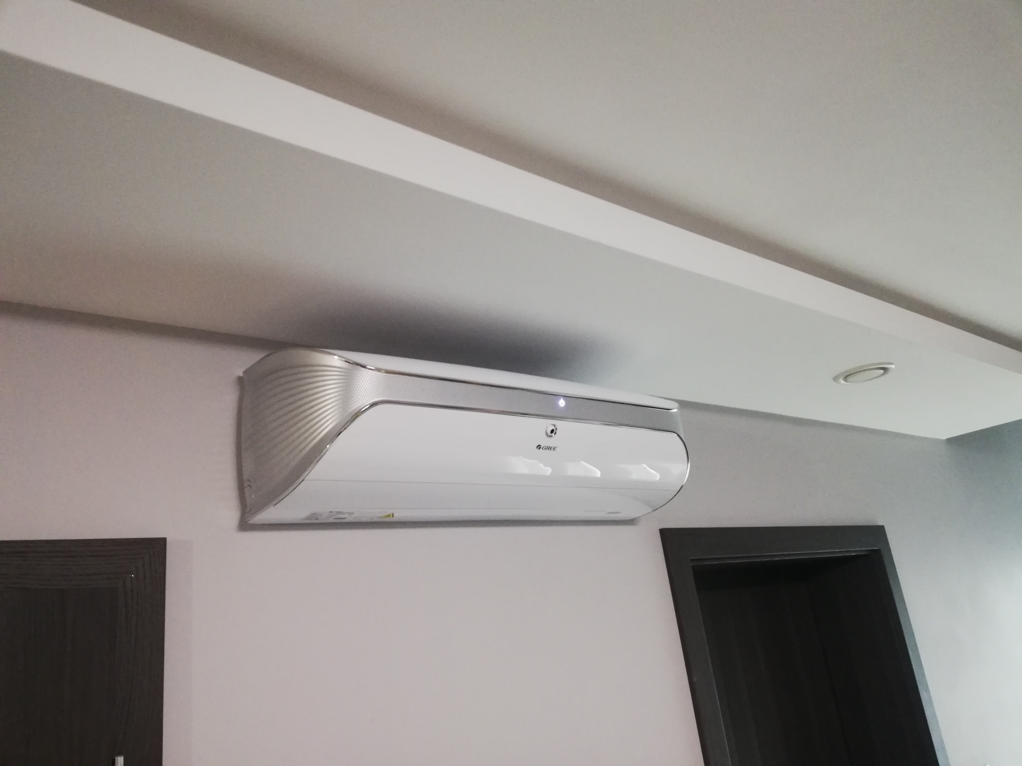Świeżo zamontowany, biały klimatyzator z srebrnymi akcentami marki Gree na szarej ścianie w nowoczesnym wnętrzu, widoczny fragment ciemnej ościeżnicy drzwiowej.