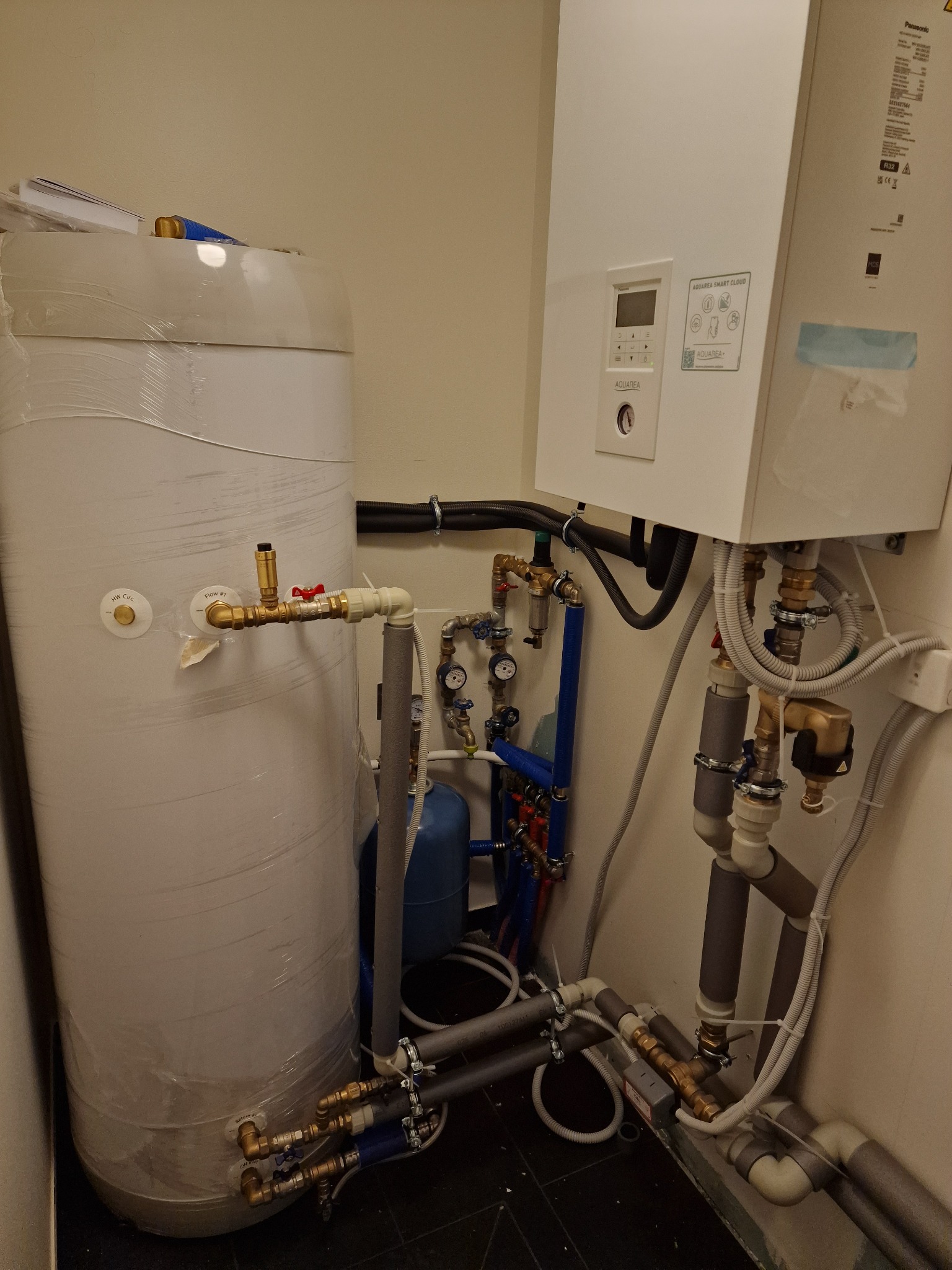 Węzeł cieplny z zasobnikiem wody i pompą ciepła Aquarea Panasonic, widoczne liczne rury, zawory i przyłącza hydrauliczne.