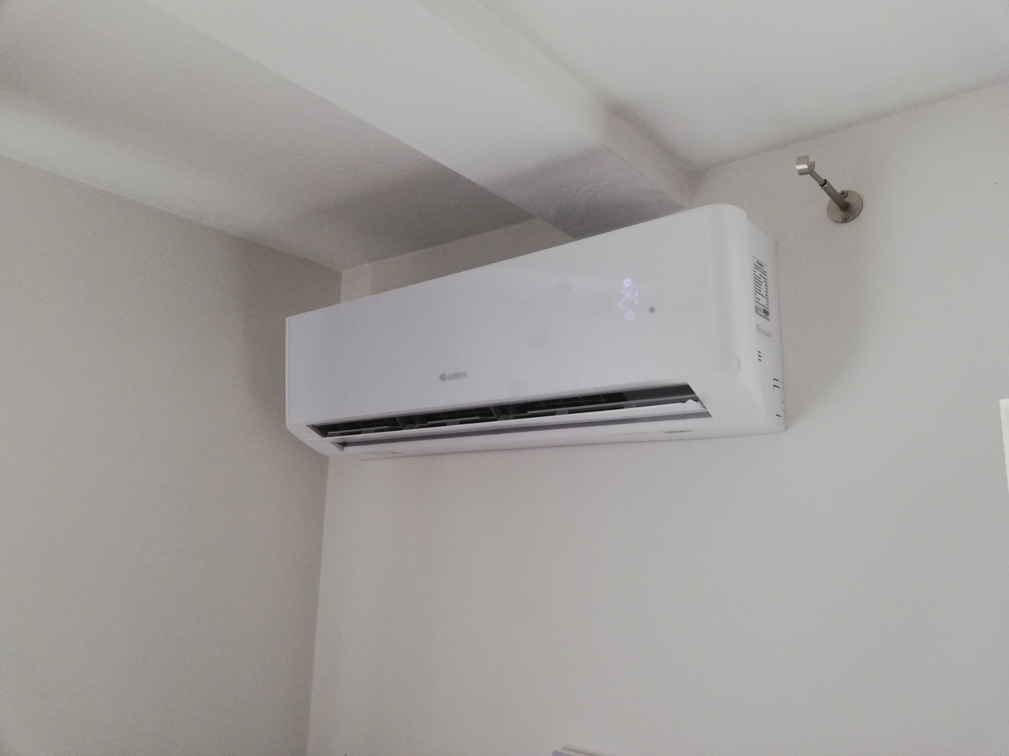 Biała klimatyzacja marki Gree zamontowana na jasnej ścianie w pomieszczeniu z widocznym fragmentem sufitu i uchwytem na karnisz.