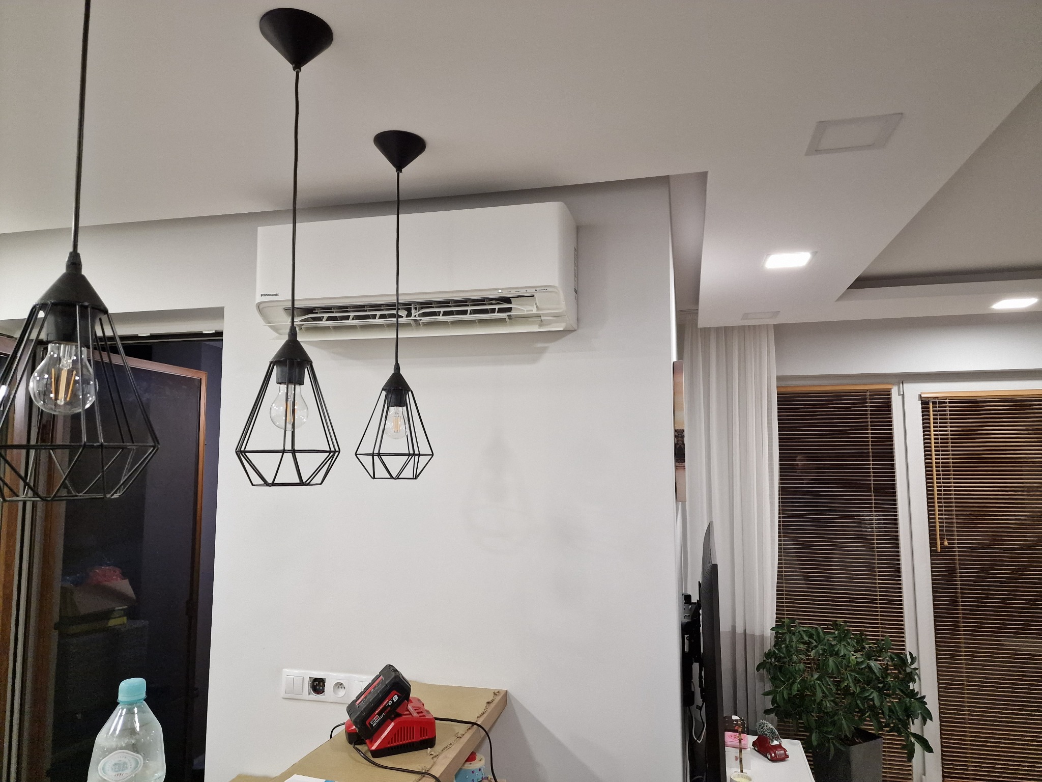 Biały klimatyzator ścienny marki Panasonic zamontowany w nowoczesnym salonie z trzema czarnymi lampami wiszącymi o geometrycznym designie, widoczne gniazdka elektryczne i ładowarka akumulatorów...