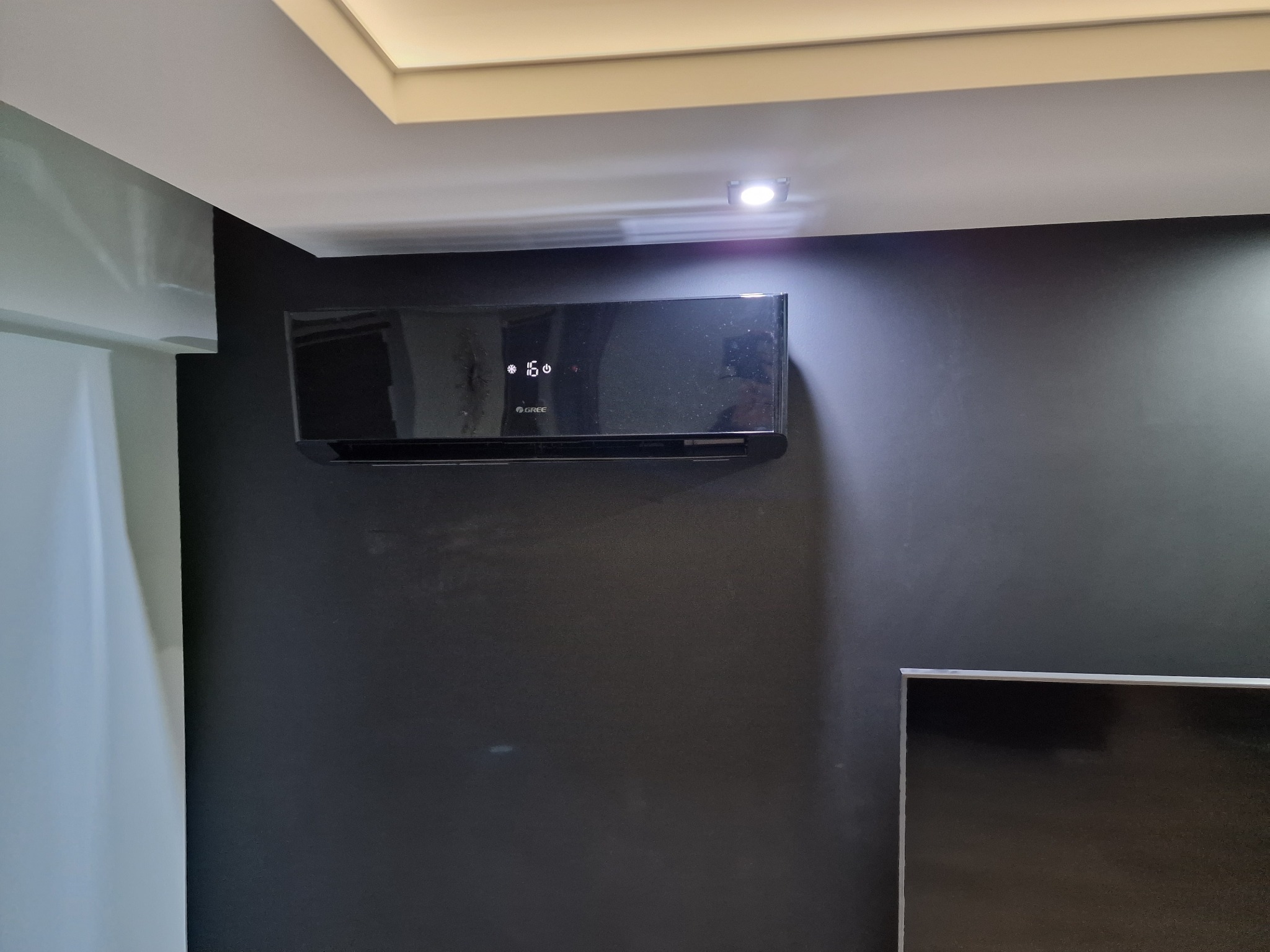 Czarny klimatyzator marki Gree zamontowany na ciemnej ścianie w pomieszczeniu z widocznym telewizorem i oświetleniem sufitowym.