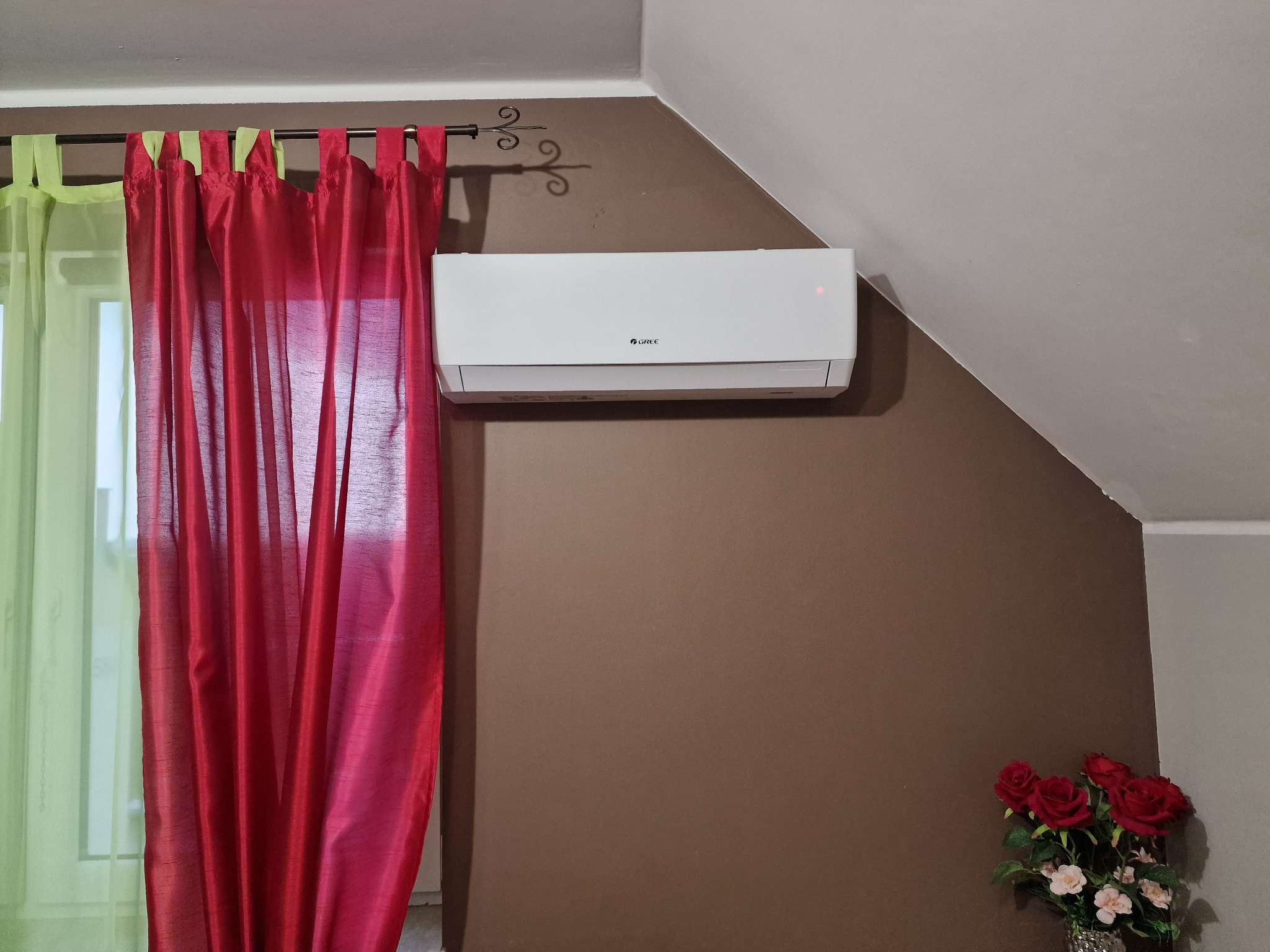 Biały klimatyzator marki Gree zamontowany na brązowej ścianie pod skosem, obok okna z czerwono-zielonymi zasłonami i bukietu czerwonych róż.