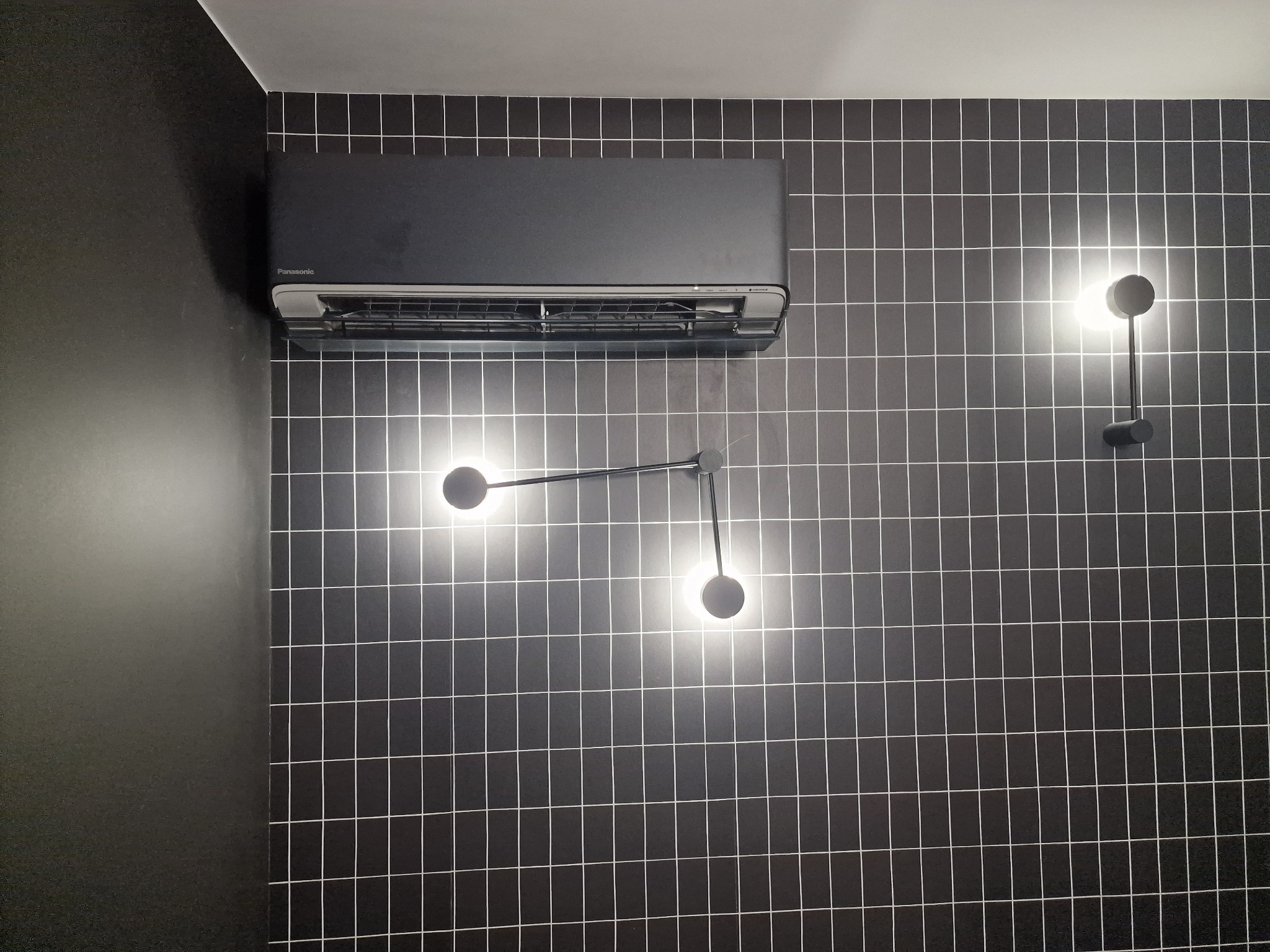 Czarna jednostka klimatyzacyjna Panasonic zamontowana na ścianie z czarnych płytek w kratkę, oświetlona trzema nowoczesnymi lampami kulistymi.