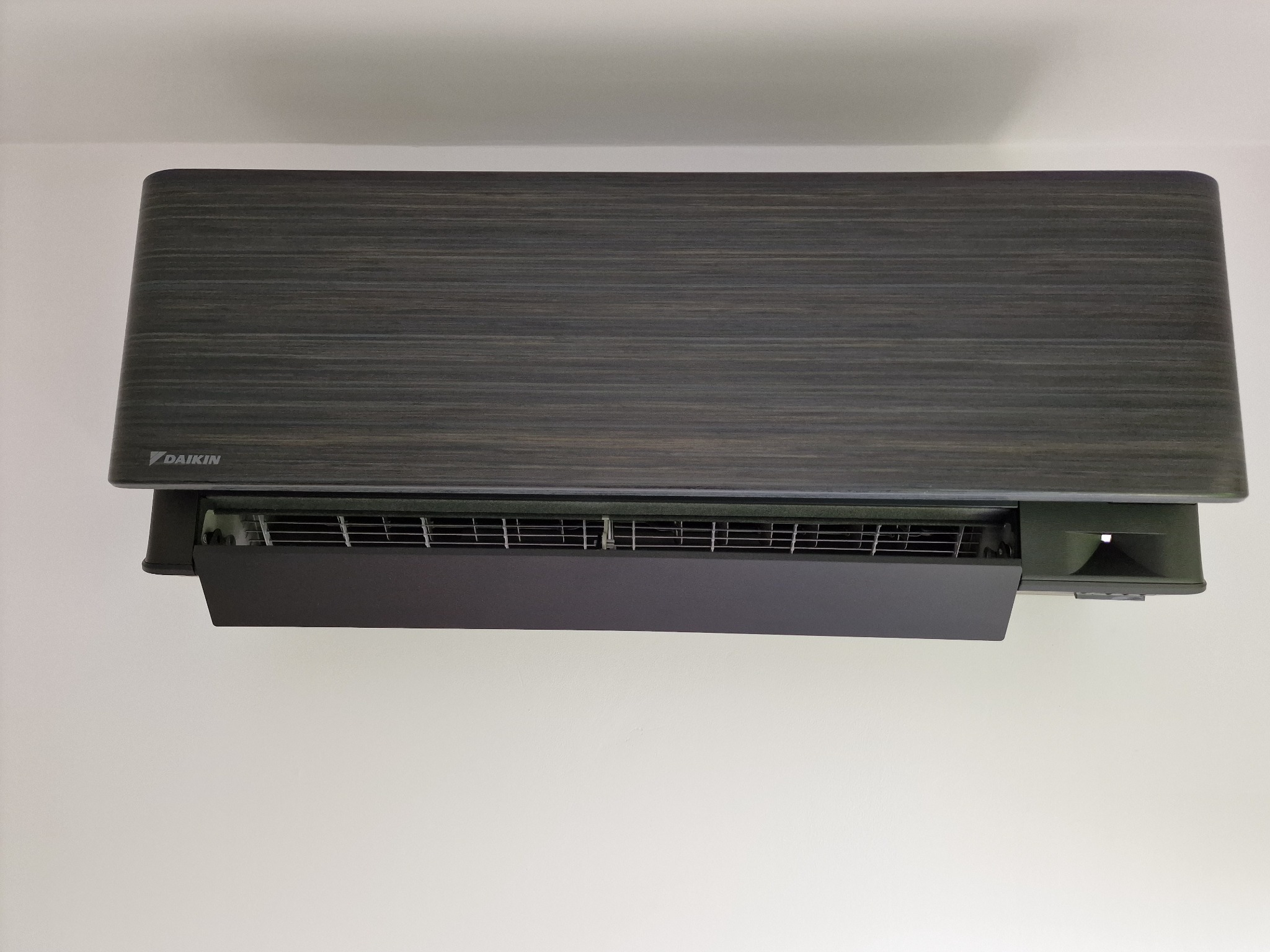 Czarny klimatyzator Daikin z panelem imitującym drewno, zamontowany na białej ścianie, widok z dołu.