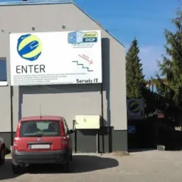 Budynek z szyldem 'ENTER Serwis IT', strzałką wskazującą wejście po schodach w dół, na parkingu stoi czerwony samochód osobowy.